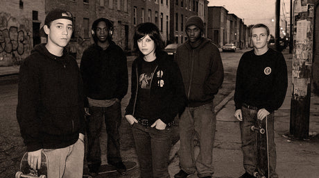 Young skaters and street culture kids on a gritty Baltimore block at dusk, early-2000s urban scene.