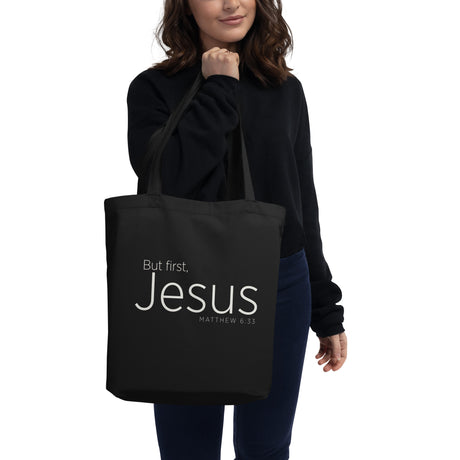 "But first, Jesus Matthew 6:33" in cream white lettering on a black Eco tote bag carried by a woman