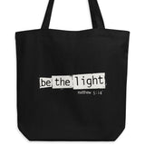 Black eco tote bag featuring a distressed cream-white “Be the Light” design with Matthew 5:14.