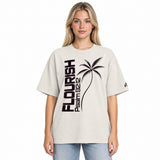 Front view of model wearing a faded bone oversized tee with dark plum vertical FLOURISH and Psalm 92:12 lettering, a palm tree graphic across the front, and a small logo on the sleeve.