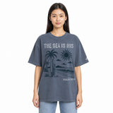 A woman modeling an oversized short-sleeve t-shirt in washed slate blue-gray. The shirt features a dark graphic print of a beach scene with palm trees, a surfboard, a sun, and waves, with the text 'THE SEA IS HIS' and 'PSALM 95:5' in white and dark gray.