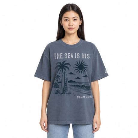 A woman modeling an oversized short-sleeve t-shirt in washed slate blue-gray. The shirt features a dark graphic print of a beach scene with palm trees, a surfboard, a sun, and waves, with the text 'THE SEA IS HIS' and 'PSALM 95:5' in white and dark gray.