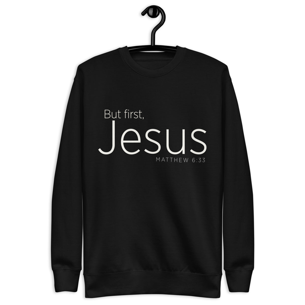 Black unisex sweatshirt with cream-white “But First, Jesus” typography and Matthew 6:33 printed on the front. Shown with hanger.