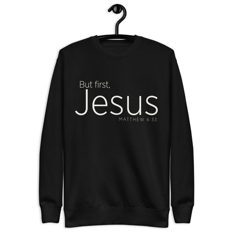 Black unisex sweatshirt with cream-white “But First, Jesus” typography and Matthew 6:33 printed on the front. Hanger view.