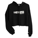 Front view of a black women’s crop hoodie with distressed white “Be The Light” lettering and Matthew 5:14 on the chest, a drawstring hood, and long sleeves.