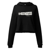 Front view of a black women’s crop hoodie with distressed white “Be The Light” lettering and Matthew 5:14 on the chest, a drawstring hood, and long sleeves.